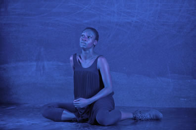 The black woman sits on the stage, wereing dark pleated tank dress over shin-length tights, her head tilted up and a tear running down her cheek